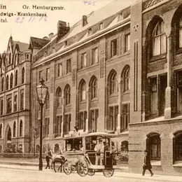 Eine historische Aufnahme des Sankt-Hedwig-Krankenhauses um 1916: Ein mehrstöckiges Gebäude in einer Häuserzeile. Vor dem Gebäude fährt ein Pferdewagen vorüber, einige Fußgänger laufen über die Straße.