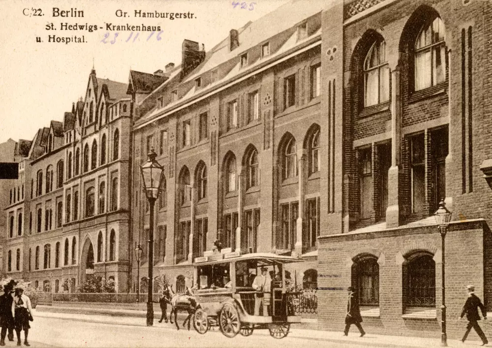 Eine historische Aufnahme des Sankt-Hedwig-Krankenhauses um 1916: Ein mehrstöckiges Gebäude in einer Häuserzeile. Vor dem Gebäude fährt ein Pferdewagen vorüber, einige Fußgänger laufen über die Straße.
