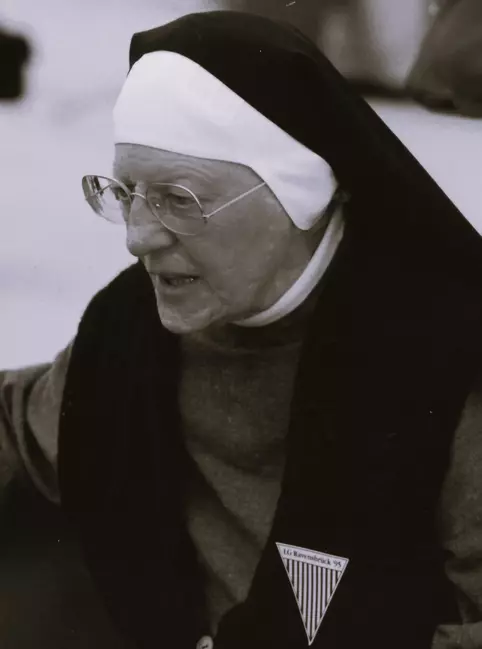 Aufnahme der 77-jährigen Katharina Katzenmaier. Sie trägt eine Ordenstracht und eine runde Brille. An ihrem Revers trägt sie ein Dreieck mit Streifenmuster, dessen Spitze nach unten zeigt. Darauf stehen die Wörter "LG Ravensbrück 95"