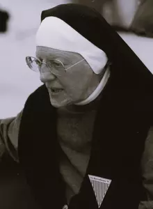 Aufnahme der 77-jährigen Katharina Katzenmaier. Sie trägt eine Ordenstracht und eine runde Brille. An ihrem Revers trägt sie ein Dreieck mit Streifenmuster, dessen Spitze nach unten zeigt. Darauf stehen die Wörter "LG Ravensbrück 95"