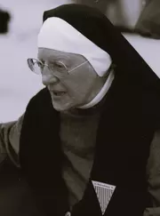 Aufnahme der 77-jährigen Katharina Katzenmaier. Sie trägt eine Ordenstracht und eine runde Brille. An ihrem Revers trägt sie ein Dreieck mit Streifenmuster, dessen Spitze nach unten zeigt. Darauf stehen die Wörter "LG Ravensbrück 95"