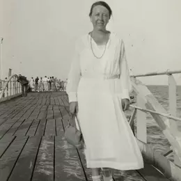 Die 40-jährige Marianne Hapig läuft über einen Steg. Sie trägt ein weißes Kleid und weiße Sandalen. In der rechten Hand trägt sie eine kleine Handtasche.
